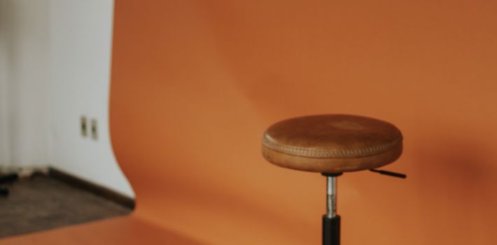 A leather stool standing empty in front of an orange studio backdrop, with a lighting stand to one side