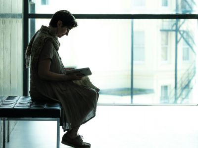 A woman sitting alone on a bench in a large, light-filled hall, reading.